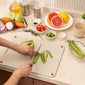Person cutting green peppers on a white cutting board with various fruits and vegetables on a kitchen counter.